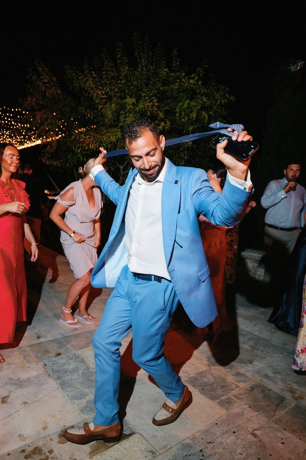 Man in light blue suit dancing enthusiastically at outdoor celebration under string lights.