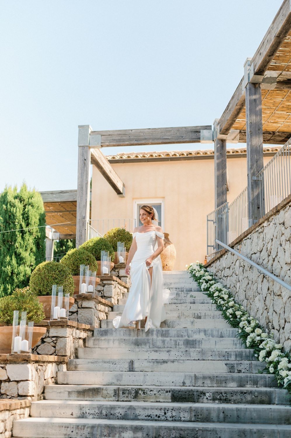 A figure in a flowing white dress descends stone steps decorated with lanterns at a Mediterranean villa.