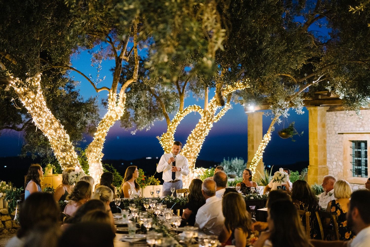 Evening wedding celebration under trees wrapped in twinkling lights against a twilight sky.