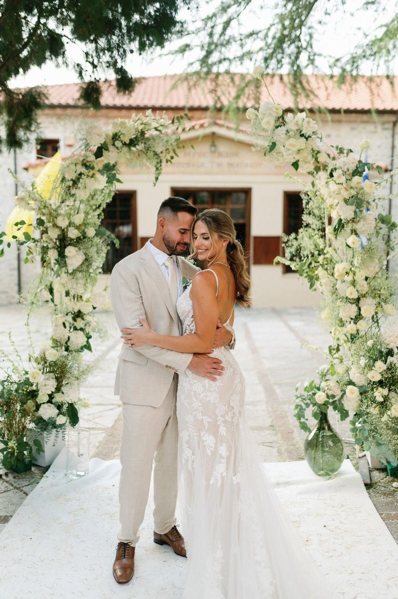 Romantic embrace under floral wedding arch with Mediterranean building backdrop.