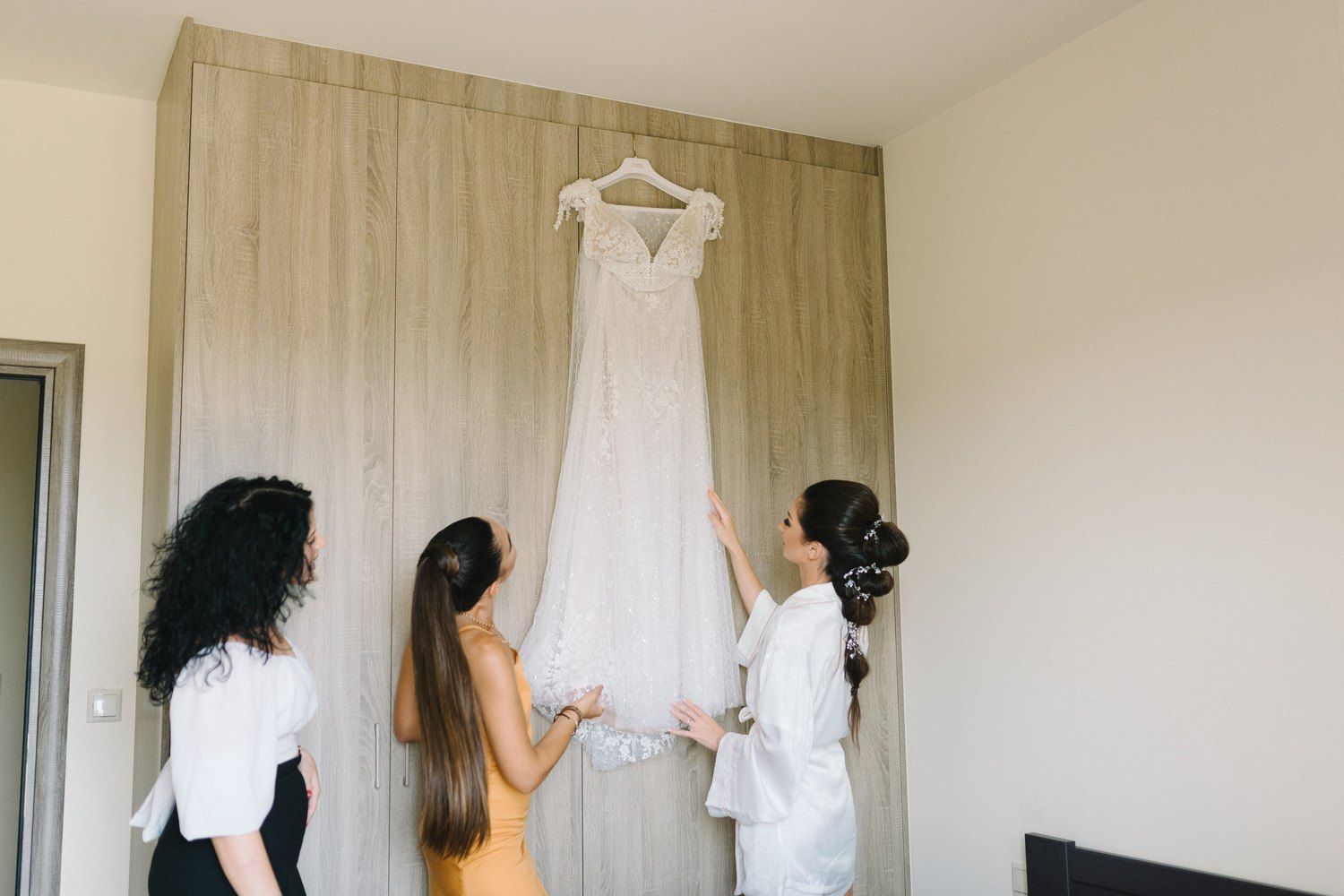 Wedding party admiring white bridal gown hanging on wooden wall.