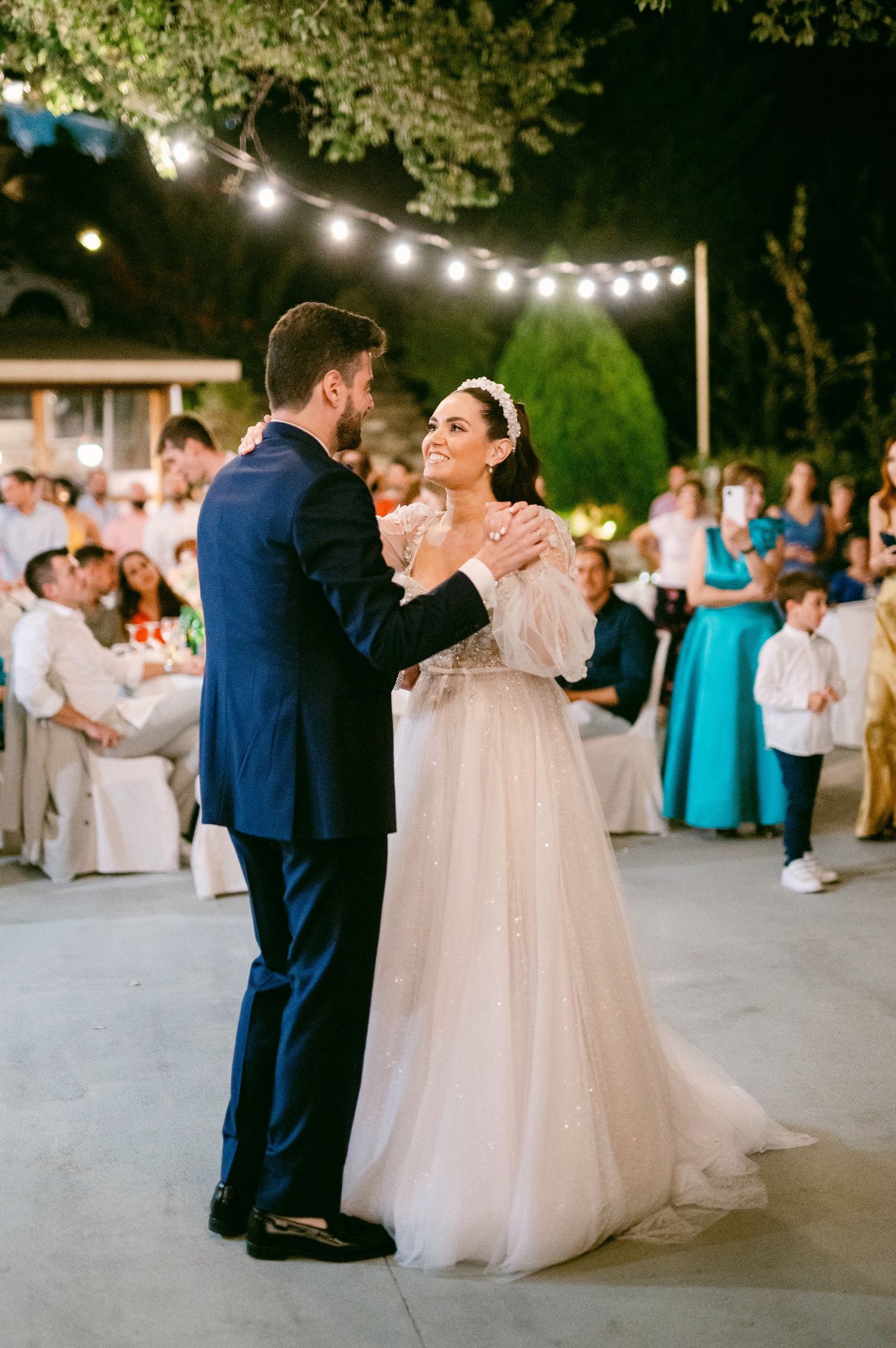 A couple shares their first dance at an outdoor evening wedding reception under twinkling string lights.