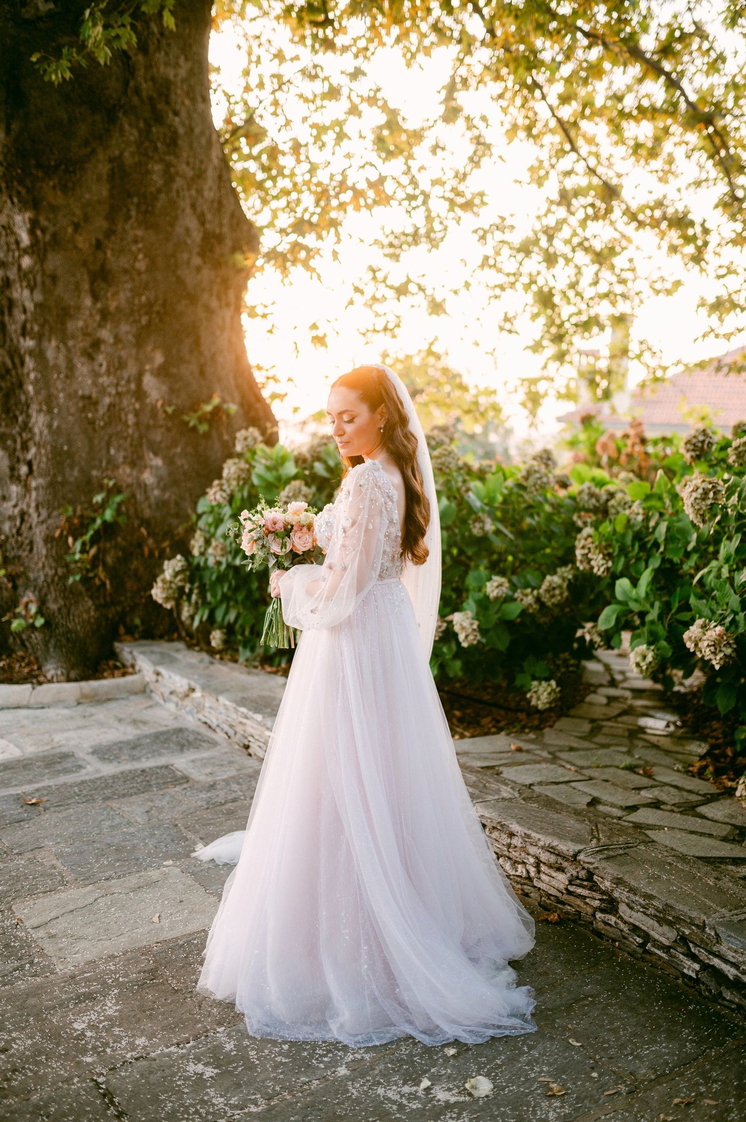 A white lace wedding gown with flowing train captured at sunset against flowering bushes and a stone wall.