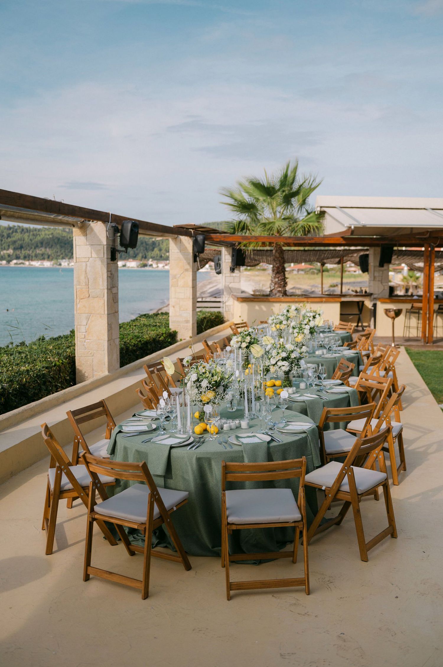 Elegant outdoor wedding reception setup with sage green tablecloths and wooden chairs overlooking a scenic waterfront view.