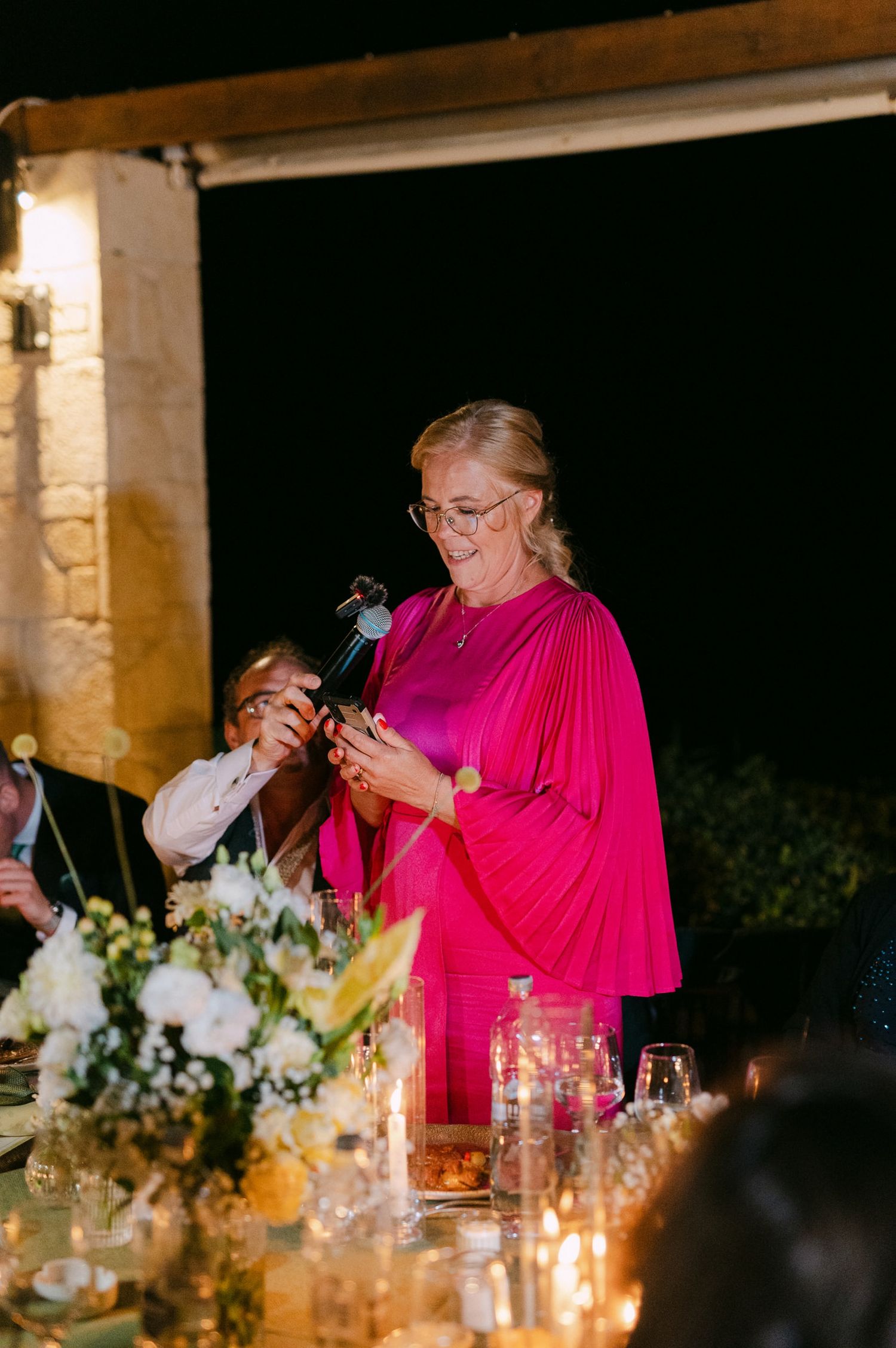 Someone in a vibrant pink dress gives a speech at an elegant candlelit dinner reception with floral centerpieces.