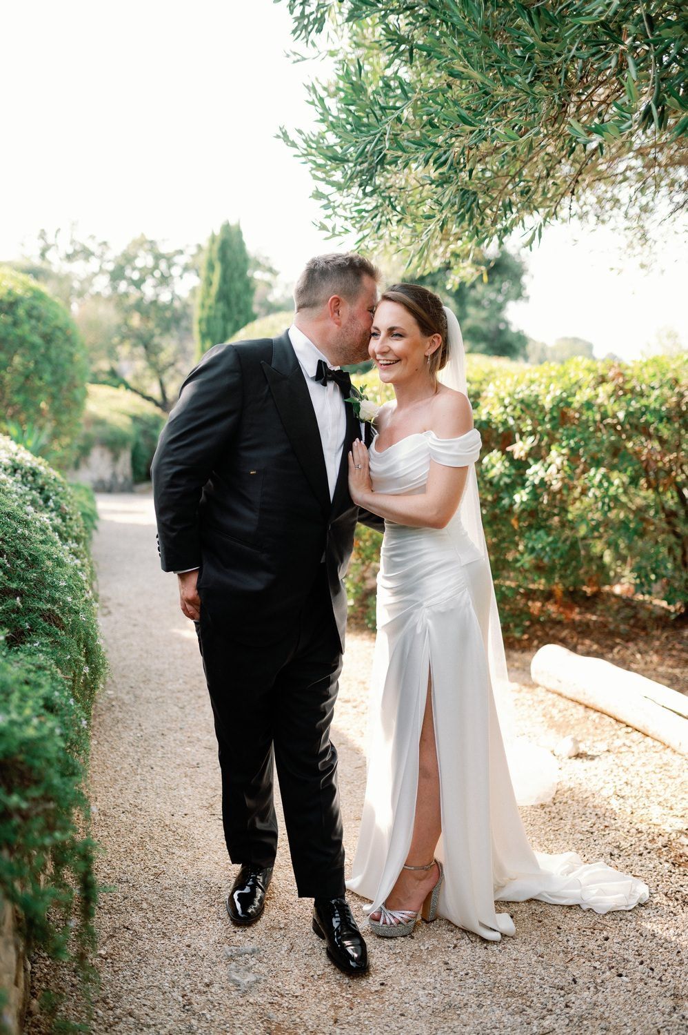 A romantic wedding couple shares tender moments on a gravel path surrounded by lush greenery and trees.