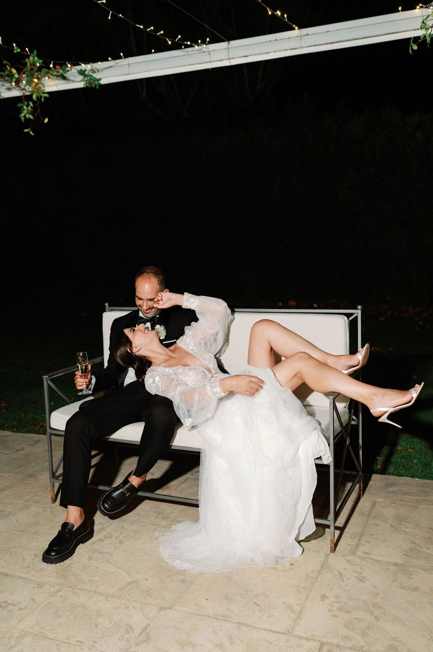 A couple relaxes on a modern outdoor bench during their wedding reception at night.