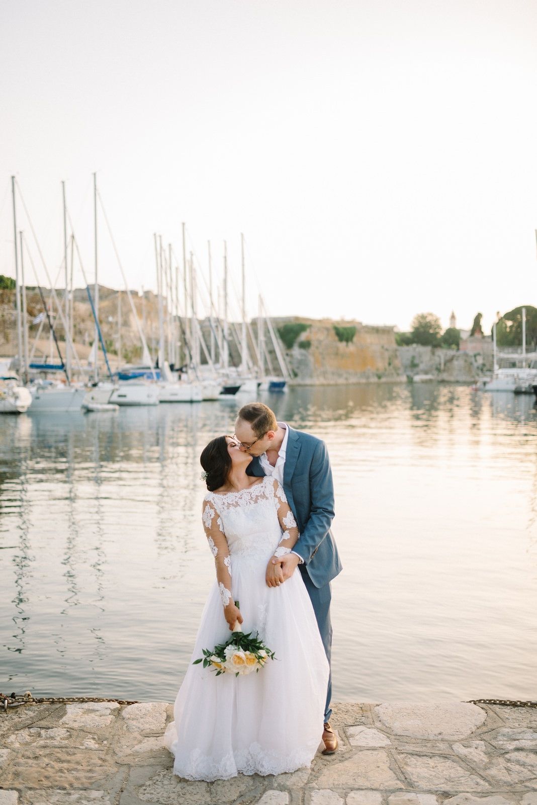 Newlyweds share a romantic moment at sunset by a marina filled with sailboats.