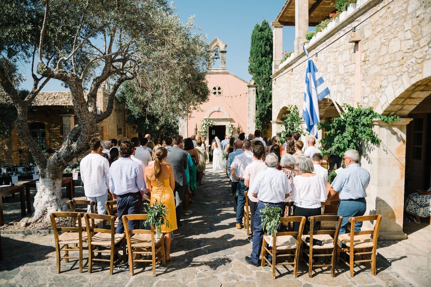 Wedding guests seated on rustic wooden chairs at an outdoor Mediterranean venue with stone walls and olive trees.