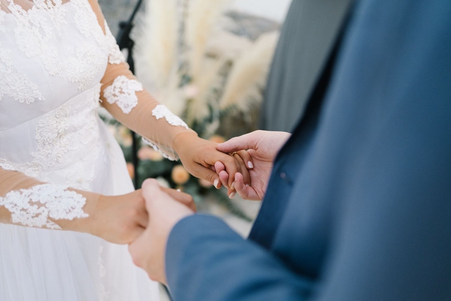 Close-up of hands exchanging rings during wedding ceremony with lace dress details.