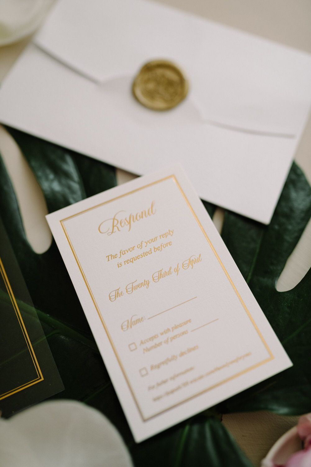 Elegant white wedding RSVP card with gold lettering and wax seal on a dark green leaf.