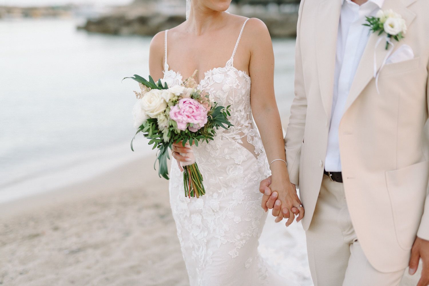 Close up of bride and groom holding hands on beach with bouquet.