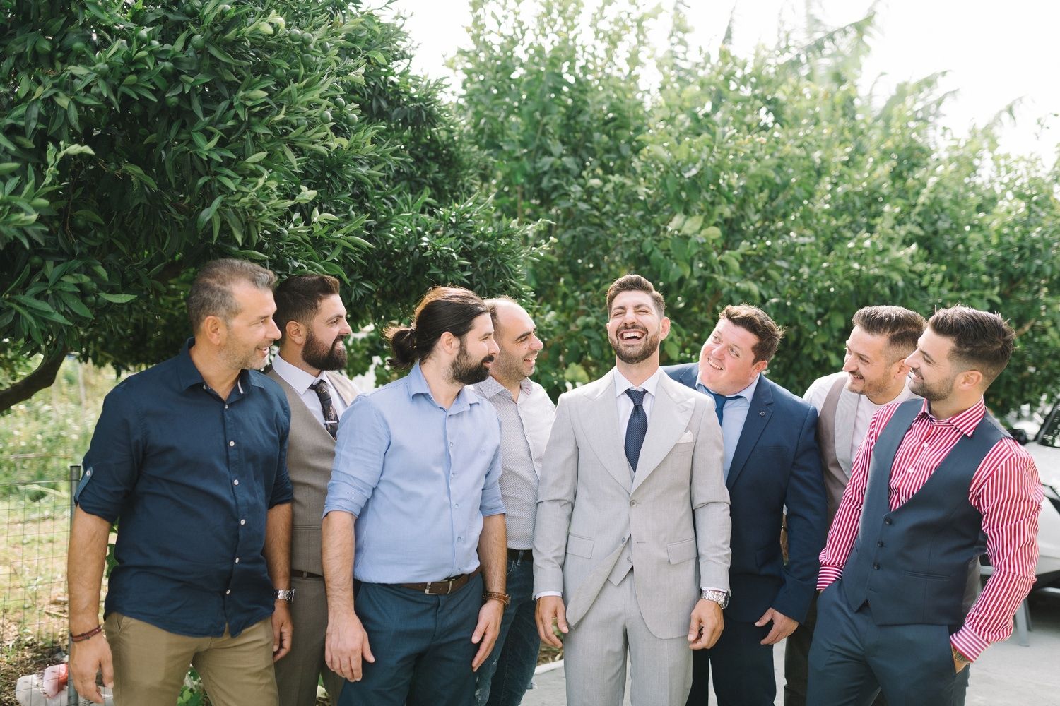 Group of groomsmen in casual and formal attire laughing together in garden setting.