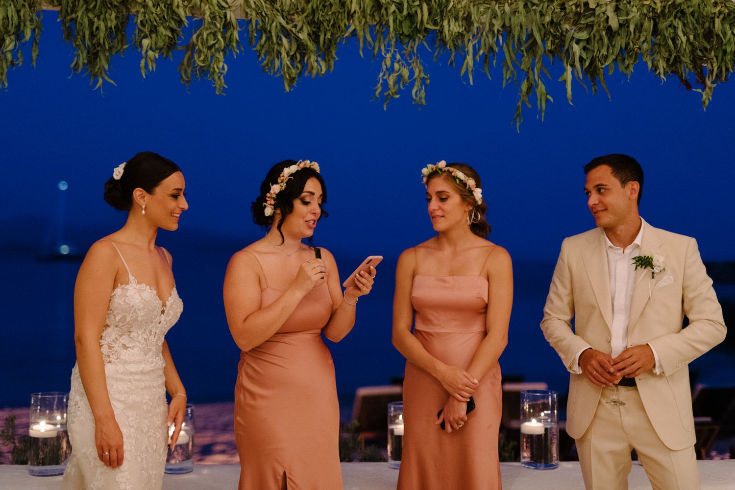Wedding party members in formal attire stand together under a canopy of lights during sunset.