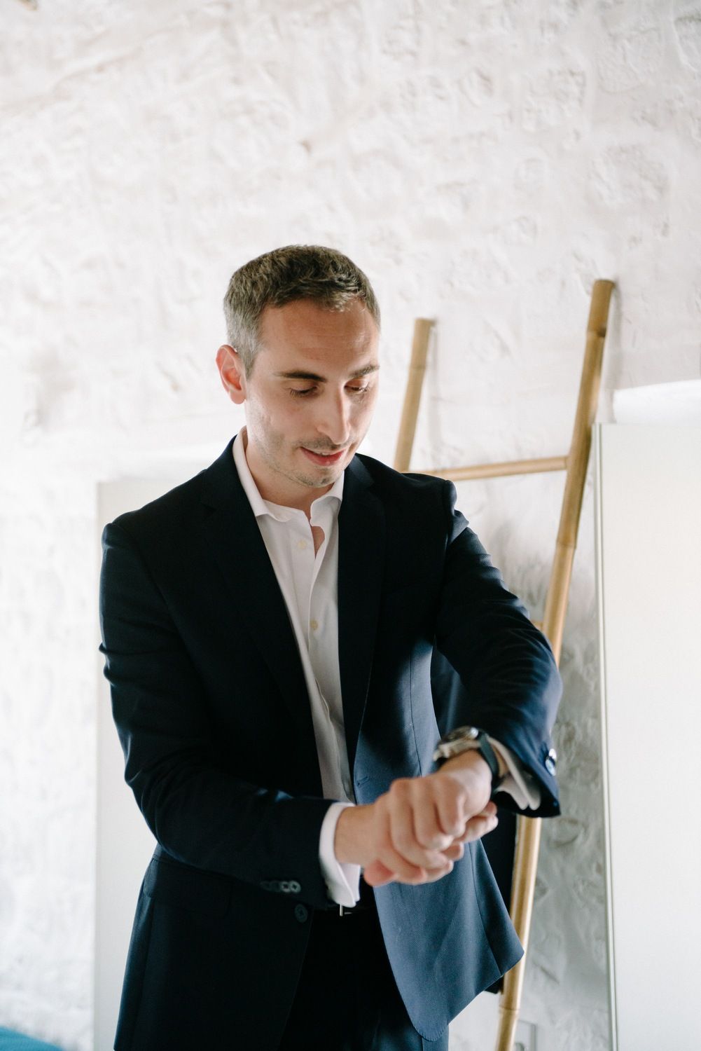 A man in a navy suit checks his watch while standing in a white-walled room.