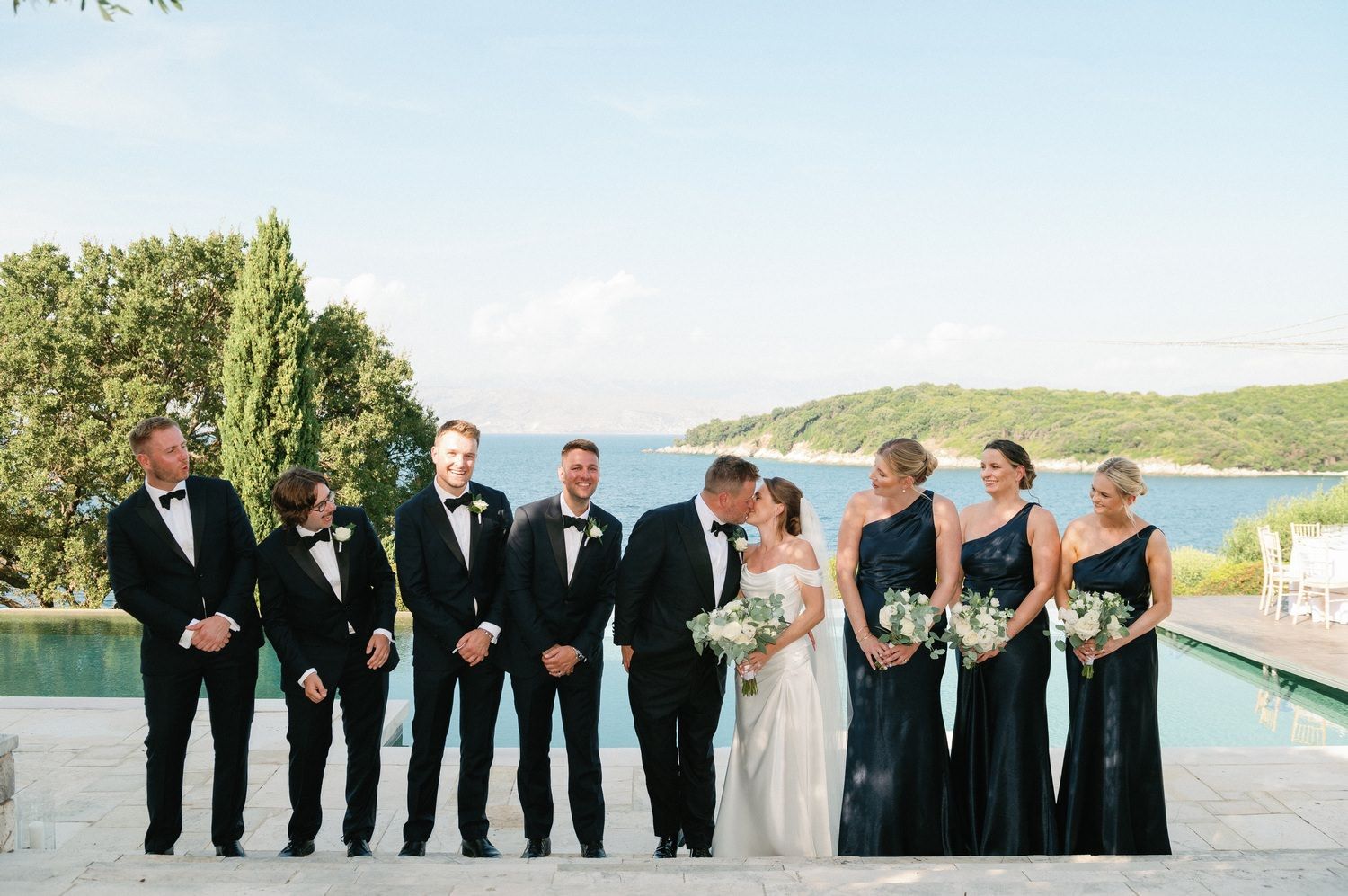 Wedding party poses against scenic coastal backdrop at an elegant outdoor venue.