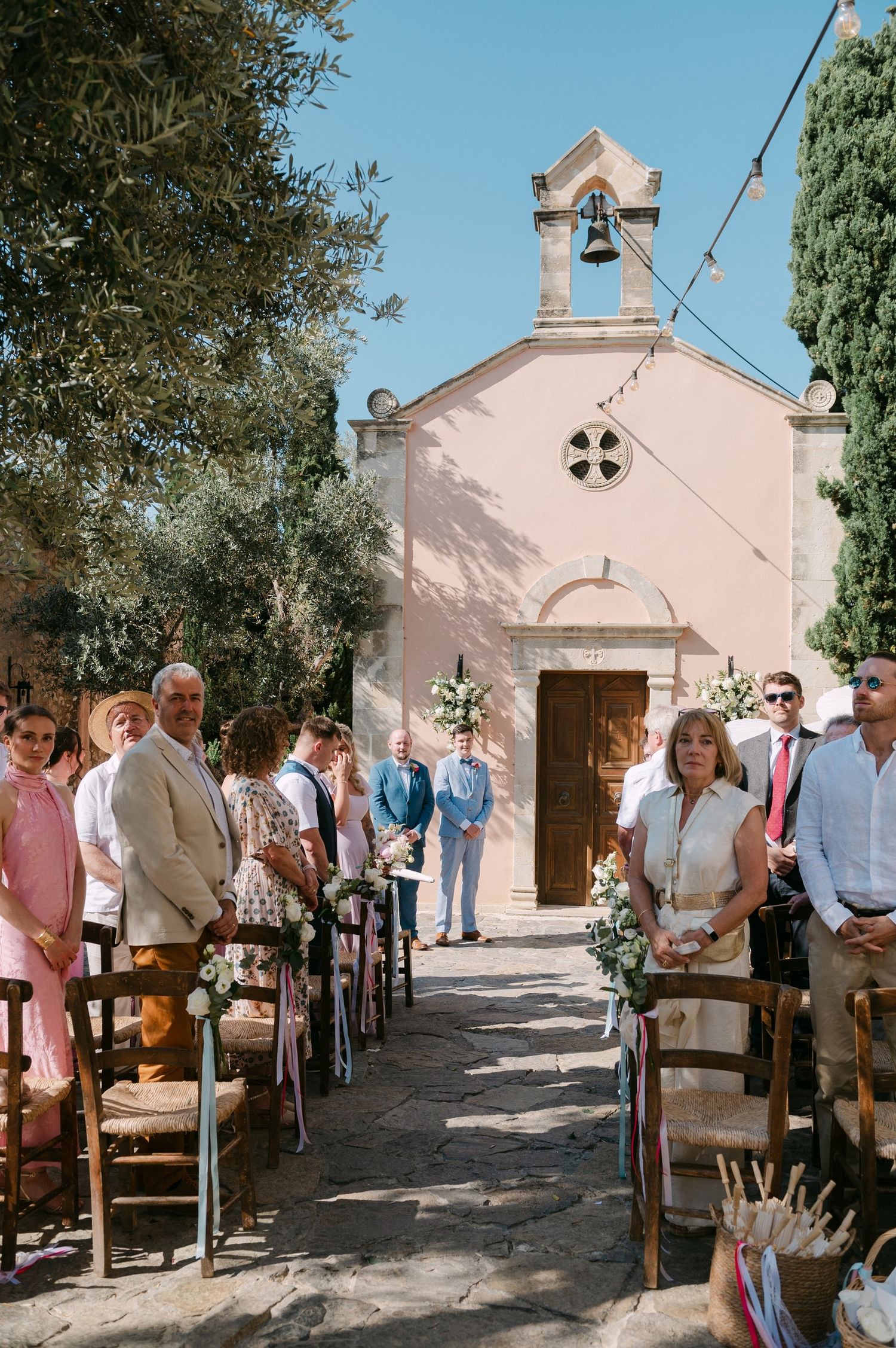 Wedding guests gather in front of a charming small pink chapel with a bell tower during an outdoor ceremony in Provence.