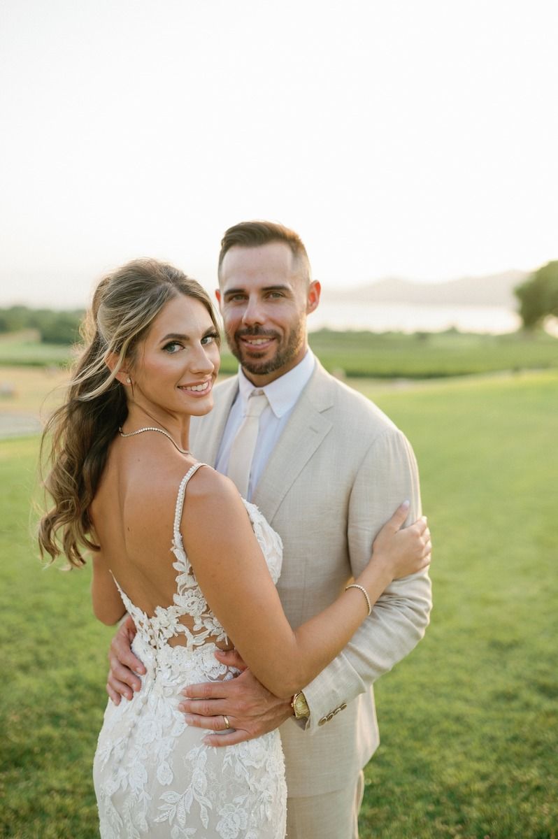 A couple embraces on a grassy hill during sunset in elegant wedding attire.