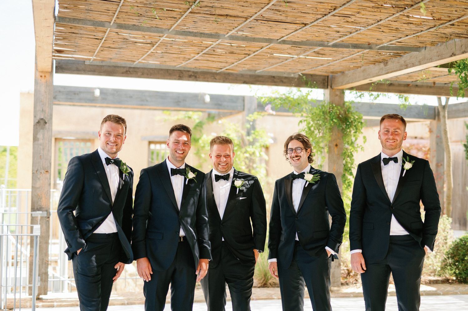 A group of groomsmen in formal black tuxedos and bow ties stand together smiling under a covered patio.