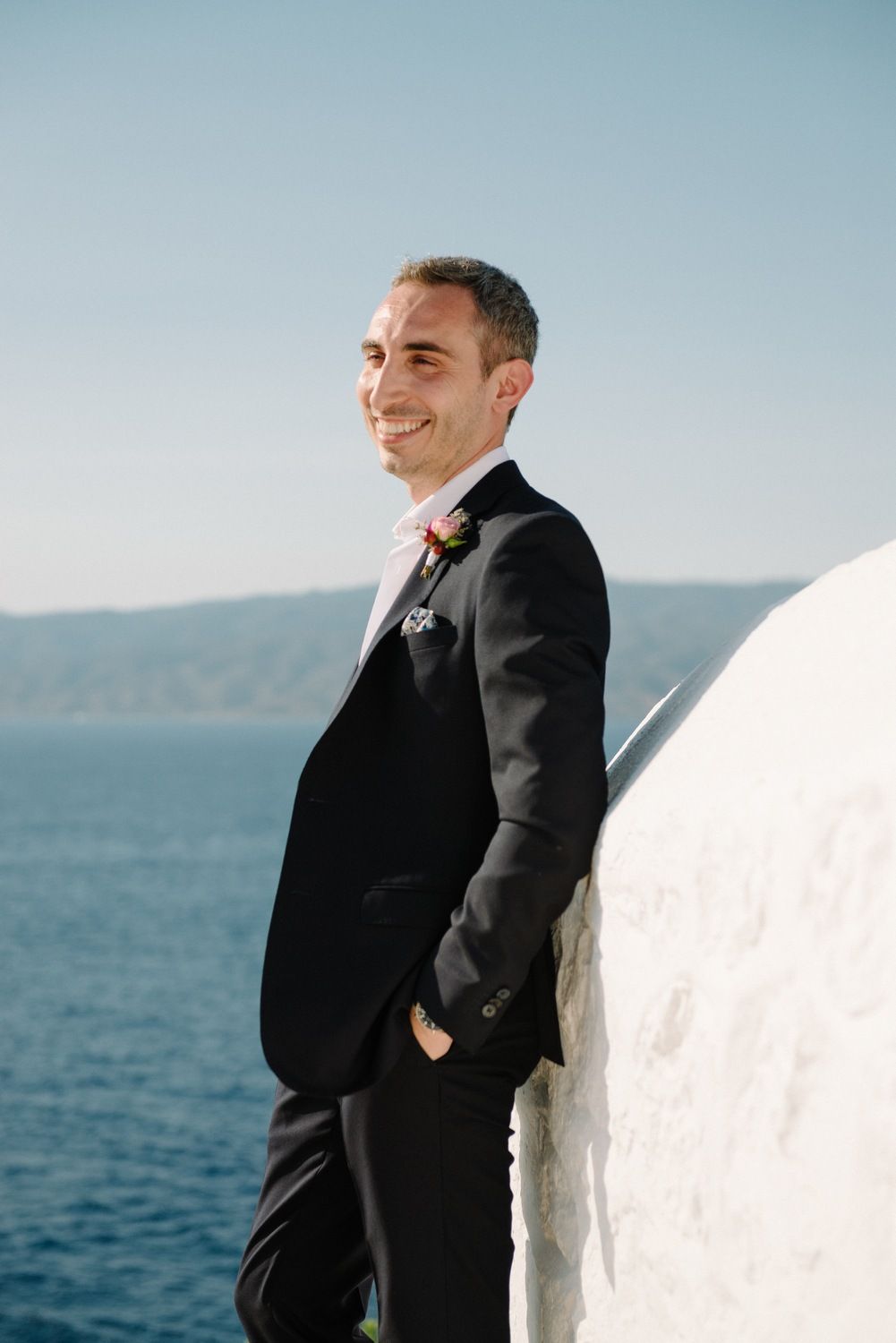 A person in dark suit smiles while standing against white Mediterranean wall overlooking ocean.