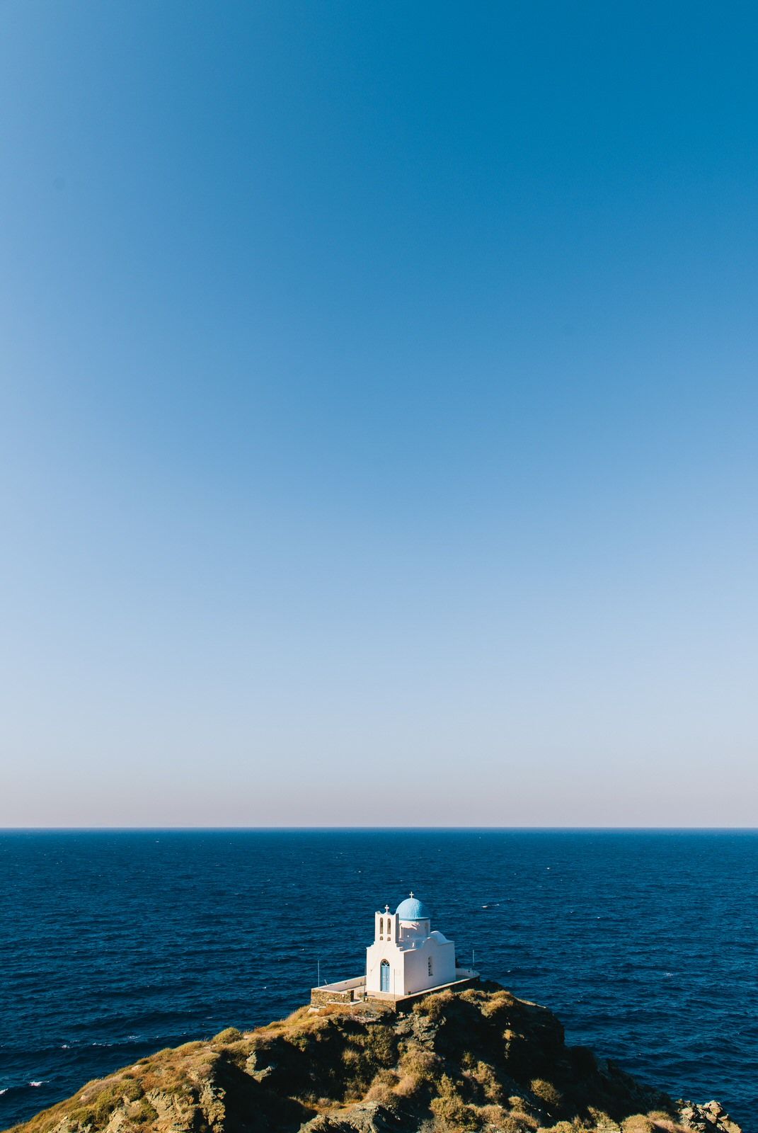 White church building perched on rocky cliff overlooking deep blue Mediterranean Sea against clear sky.