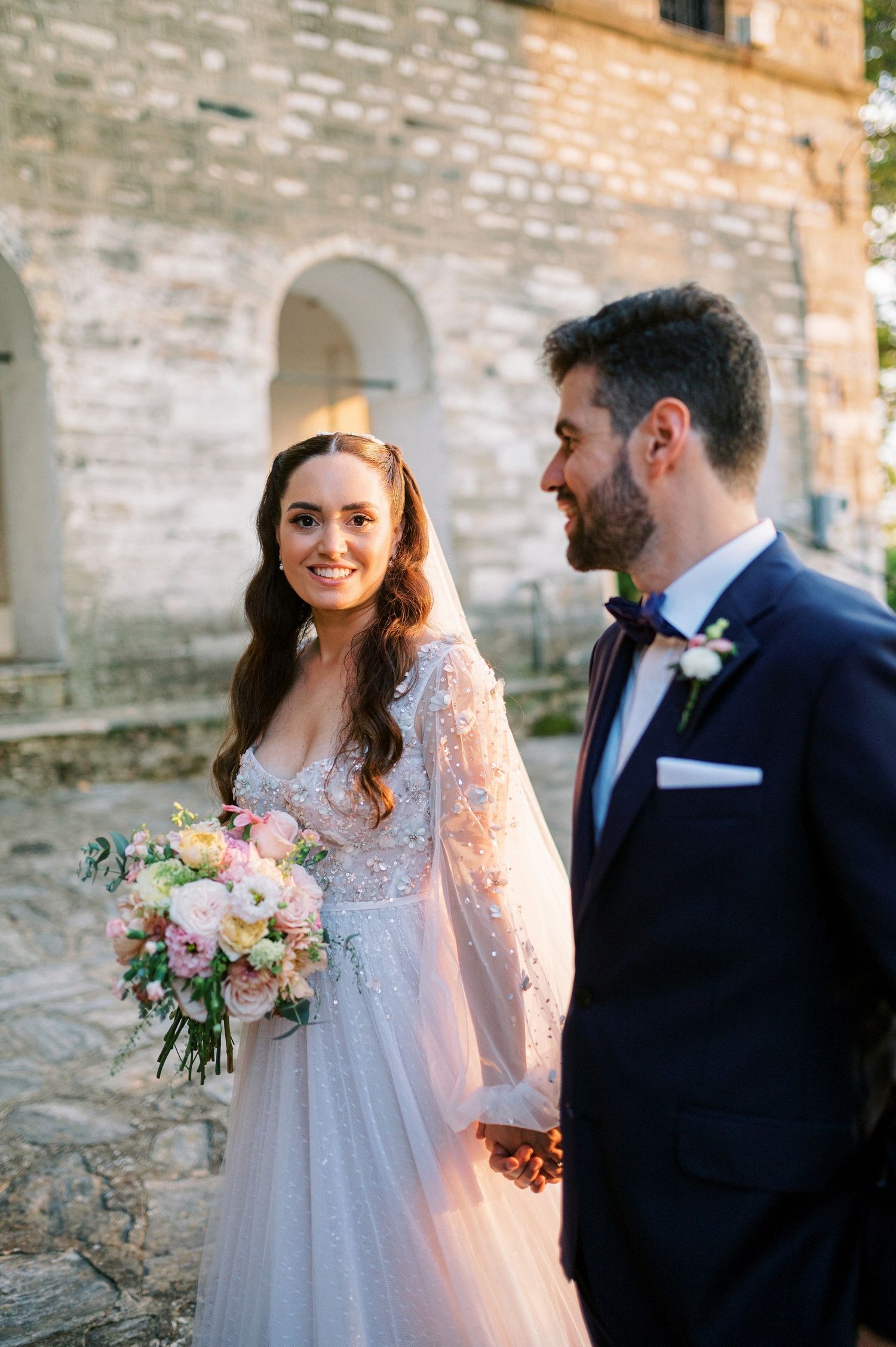 A romantic wedding couple stands together against a stone wall, with the bride holding a soft pink and peach bouquet.