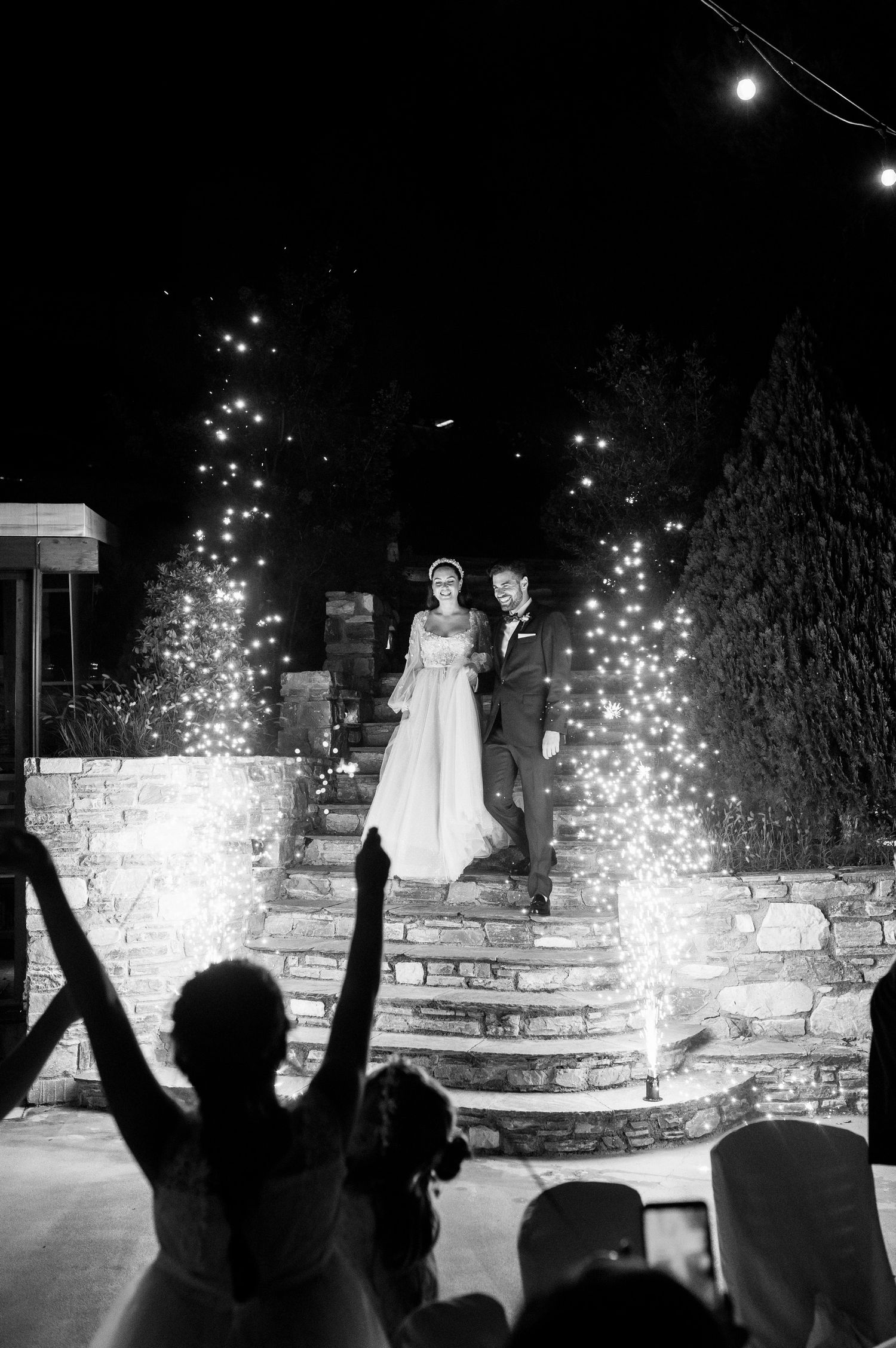 Wedding couple stands atop illuminated stone steps as silhouetted guests celebrate with sparklers at a nighttime reception.