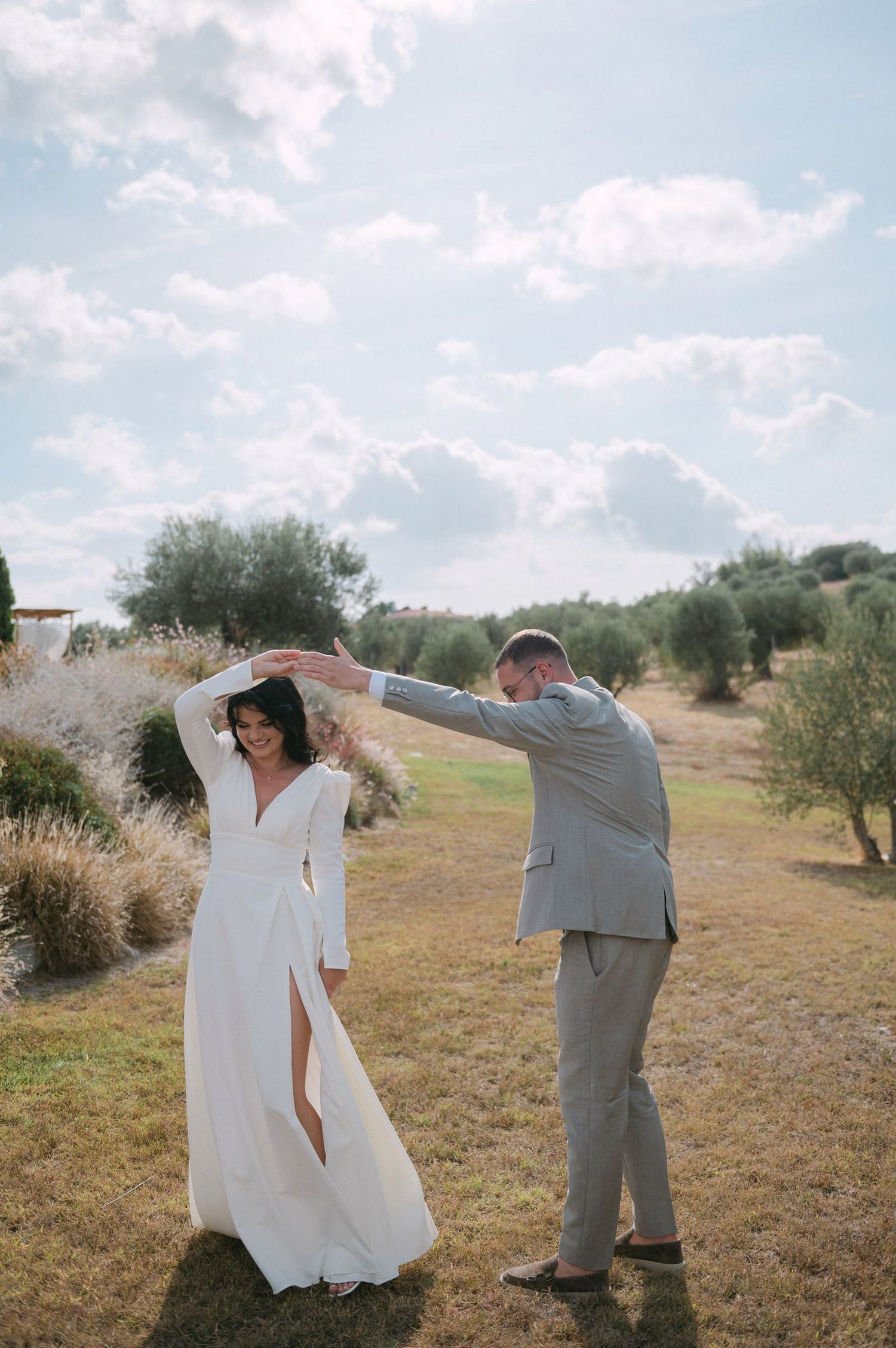A couple in wedding attire dancing together in an olive grove on a sunny day with white clouds in the sky.