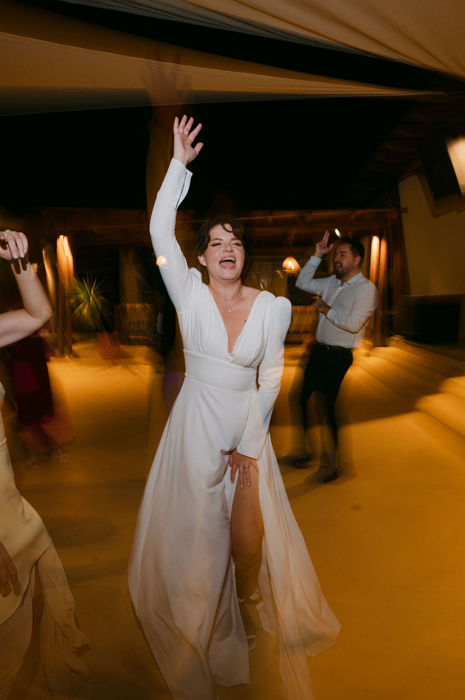 A person in a flowing white wedding dress dances joyfully on a warmly lit dance floor during a celebration.