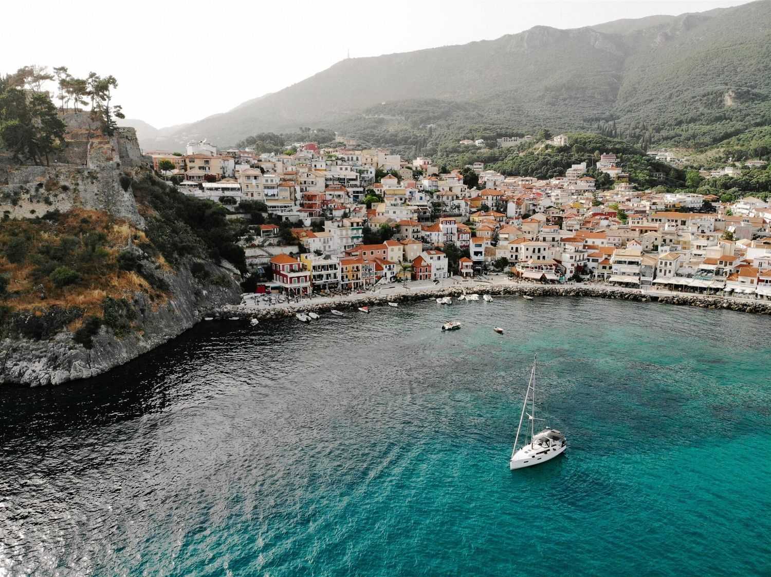 Aerial view of Parga with colorful buildings and turquoise waters featuring a white sailboat in the harbor.