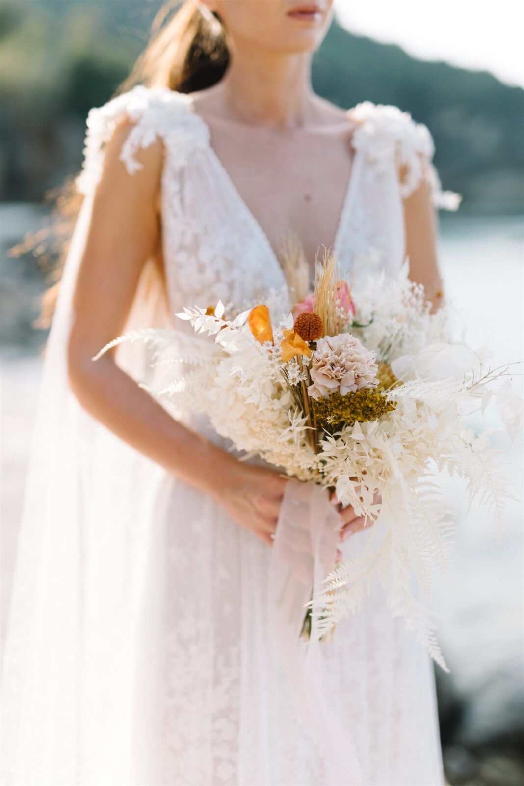 Delicate dried floral wedding bouquet with white and orange blooms held against a white lace dress.