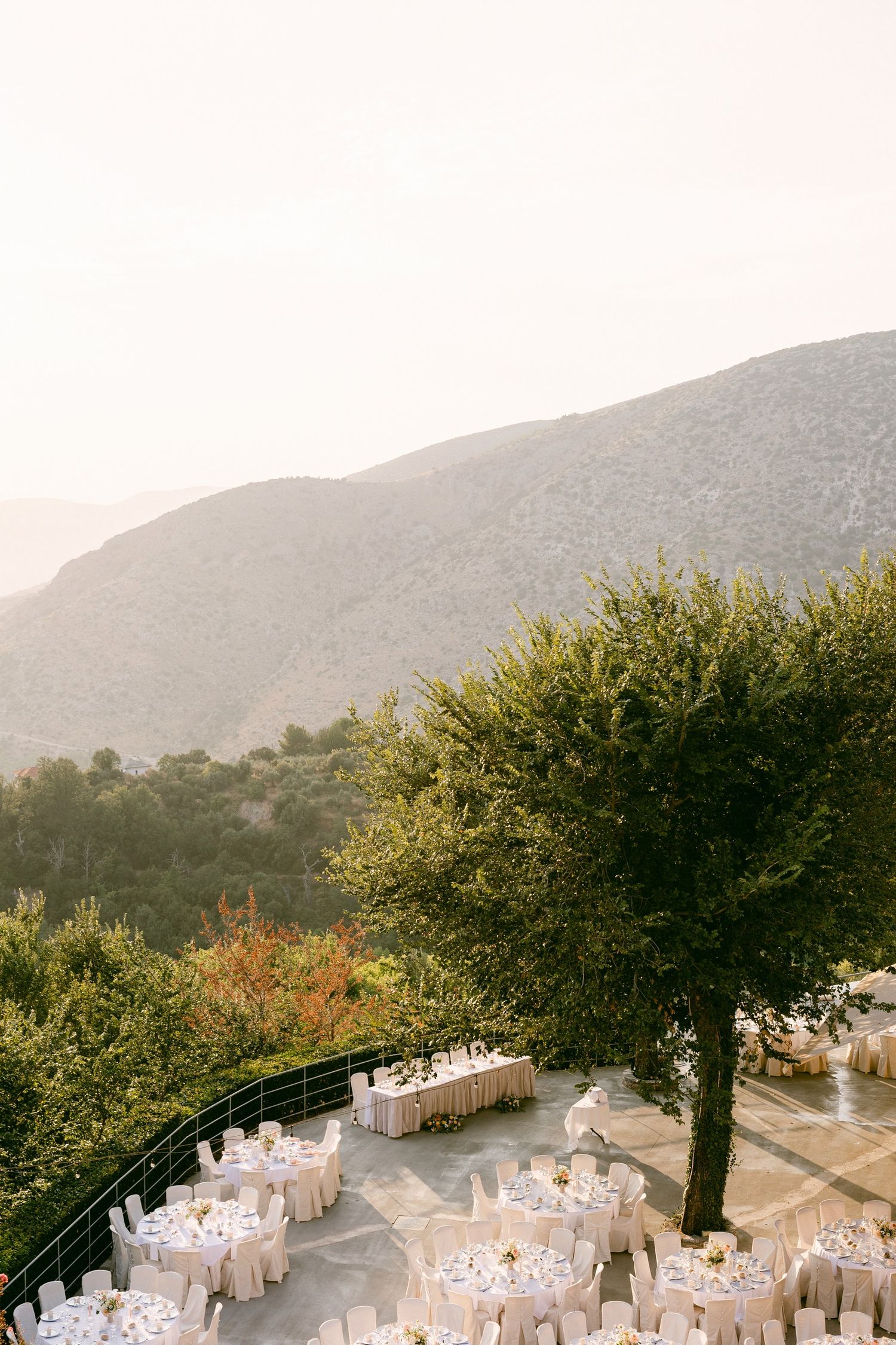 An outdoor wedding reception setup with round tables and white chairs overlooking a scenic mountainside at sunset.