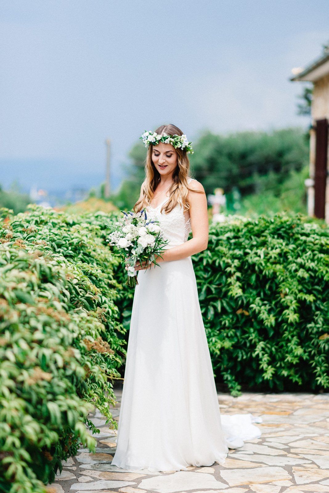 A bride in a white dress and floral crown stands on a stone patio surrounded by lush greenery and mountains.