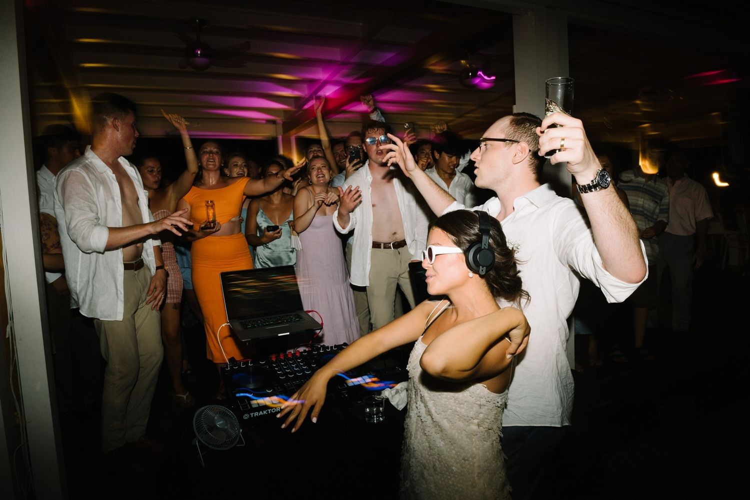 Wedding celebration on dance floor with guests enjoying music in purple lighting.