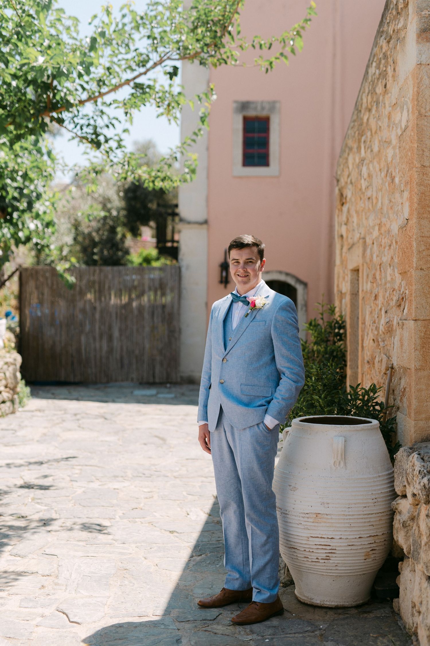 Man in light blue suit standing against pink wall with rustic clay pot in a Mediterranean garden setting.