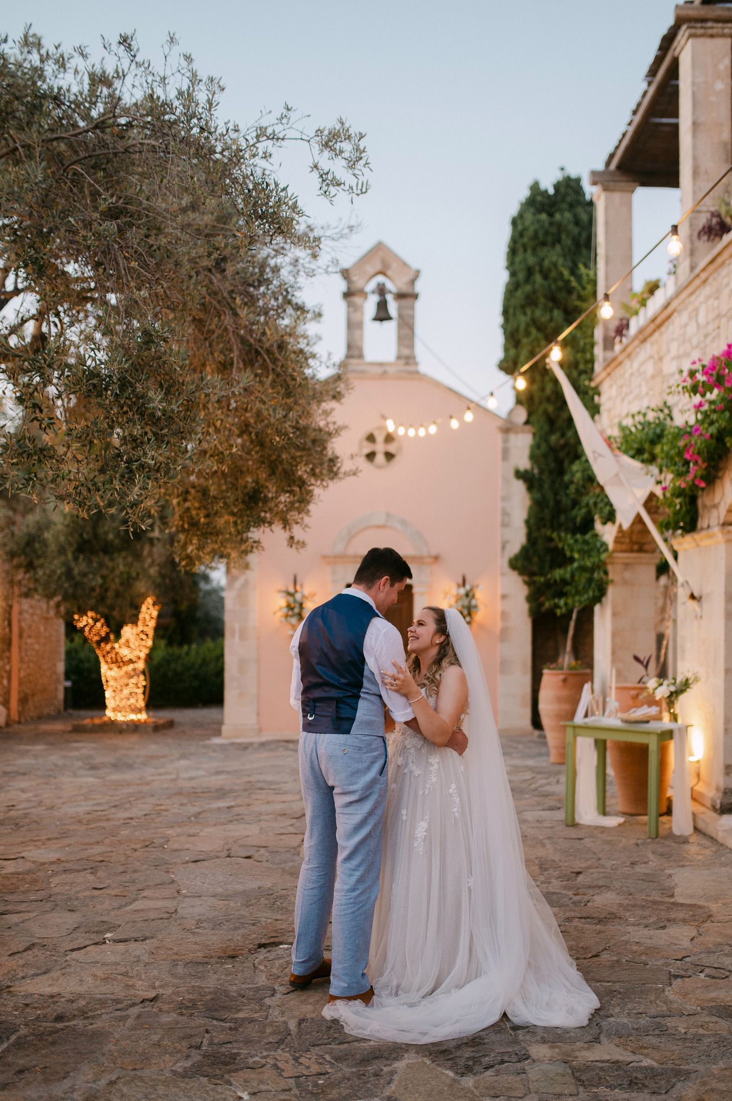 A romantic wedding couple shares an intimate moment in front of a pink Mediterranean church at sunset.