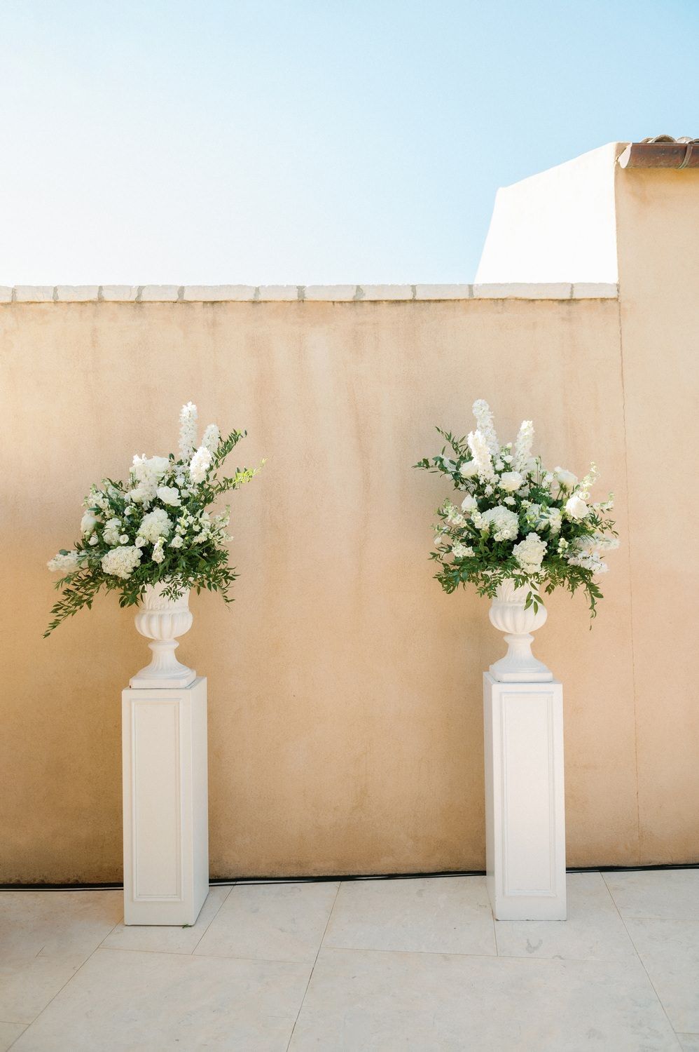 Two white floral arrangements in classic urns displayed on pedestals against a beige wall.