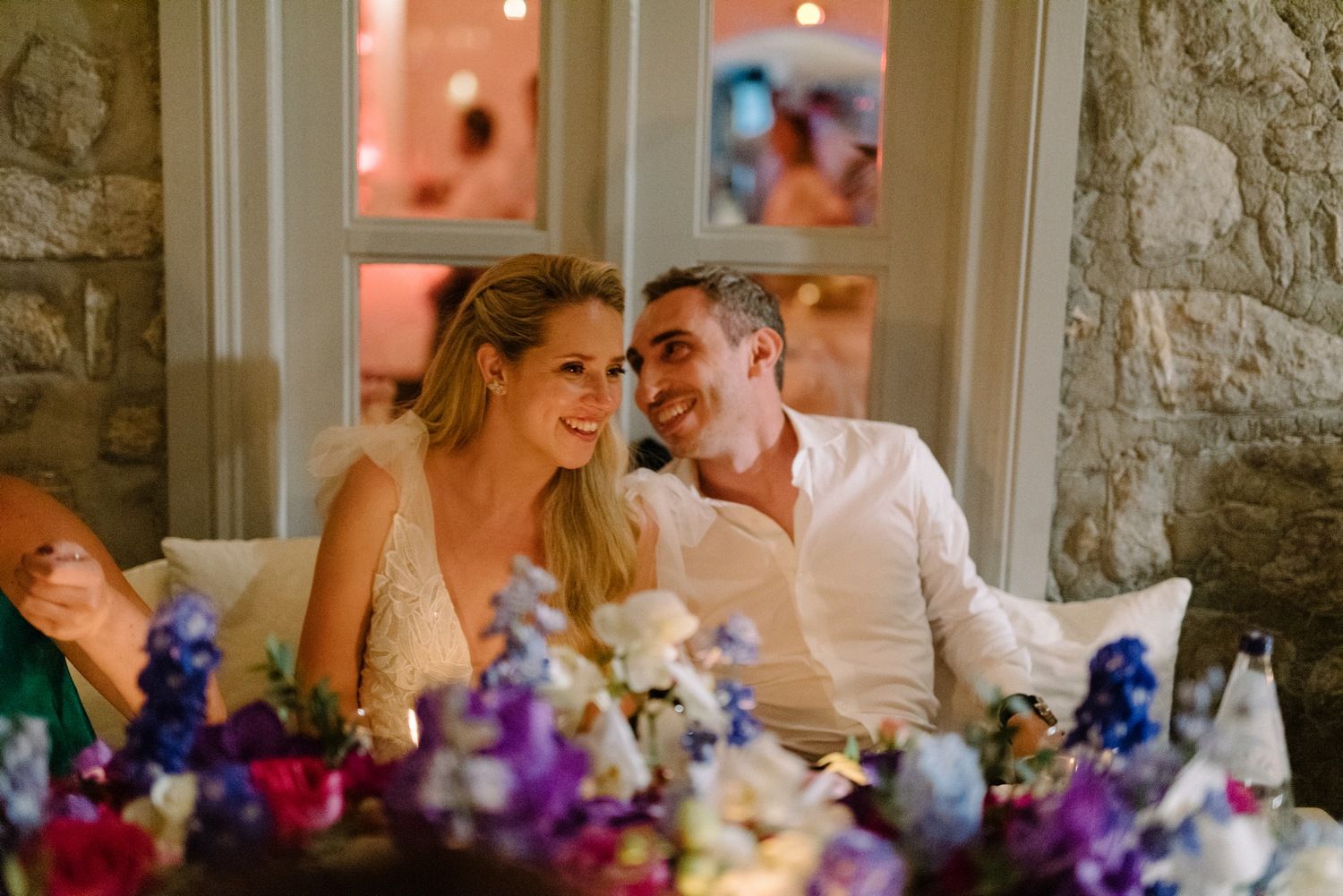 Wedding couple seated at reception table with vibrant purple and blue floral arrangements.