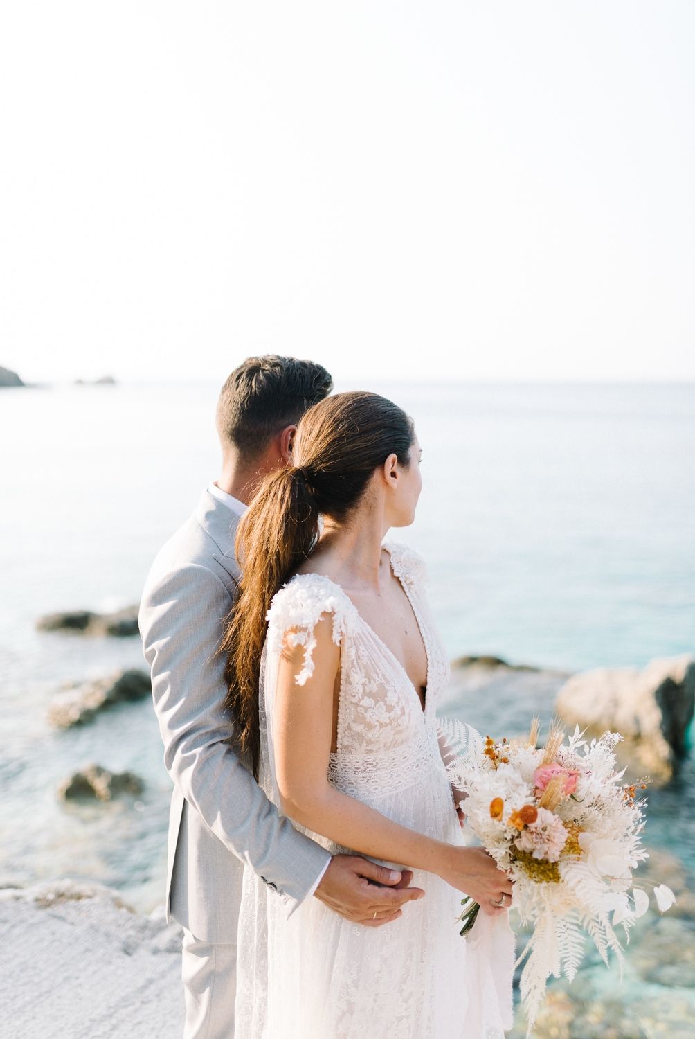 Intimate wedding portrait showing couple embracing while holding pastel bouquet by the ocean.