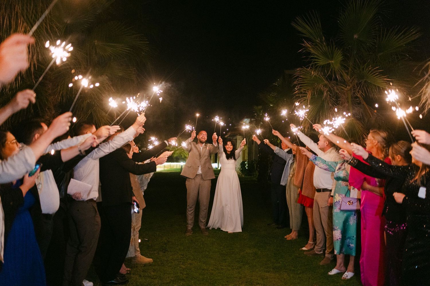 Wedding guests hold sparklers in a line at night creating a magical tunnel of light for the newlyweds.