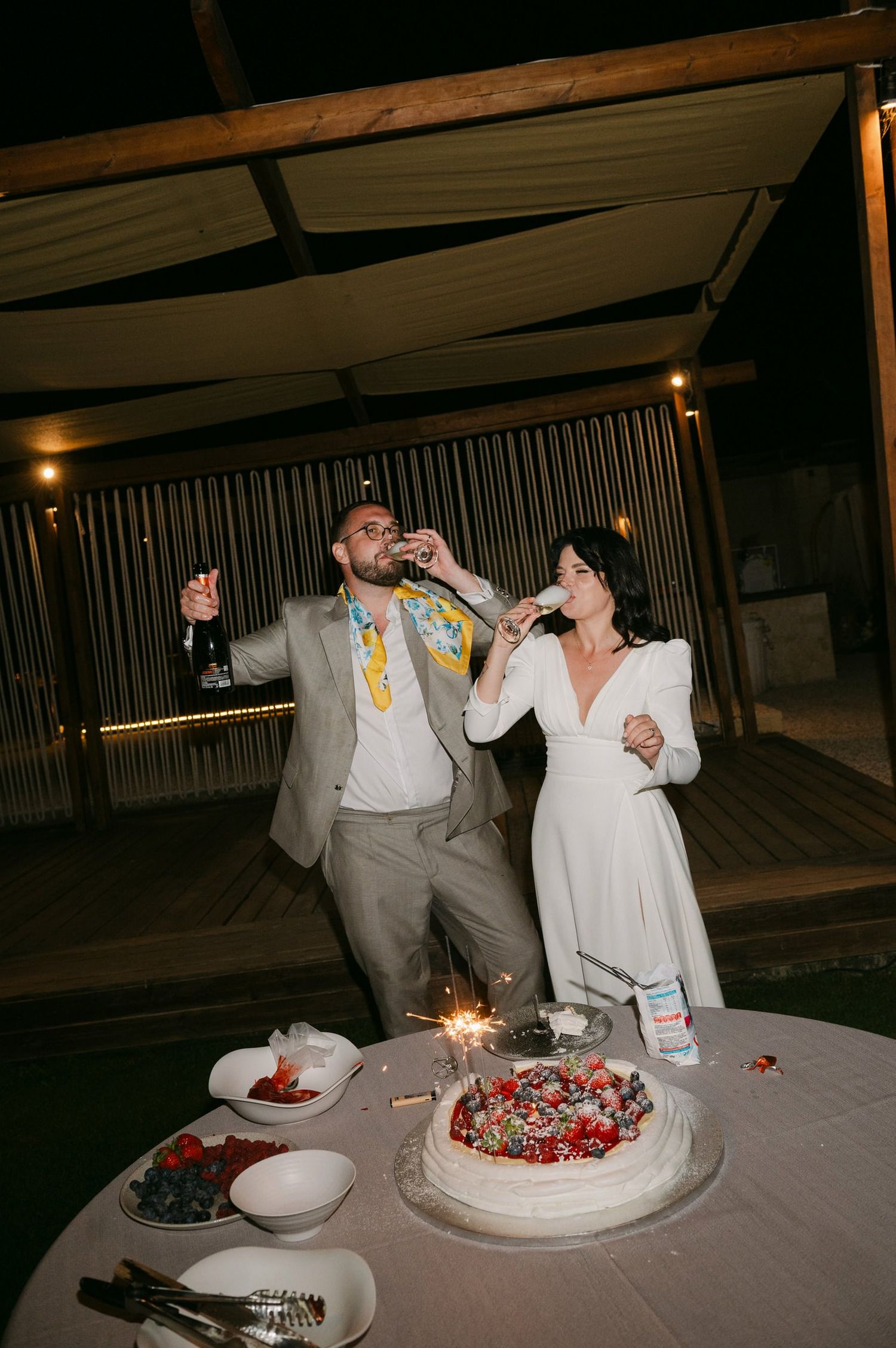 Wedding guests enjoy cake and drinks at a dimly lit reception with sparklers illuminating the festive dessert table.