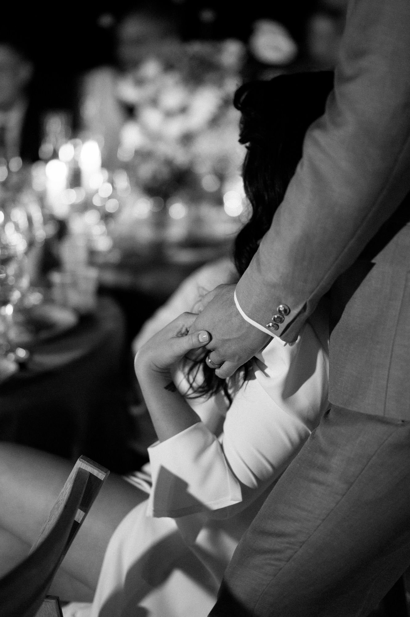 A black and white close-up of newlyweds holding hands at their wedding reception table.