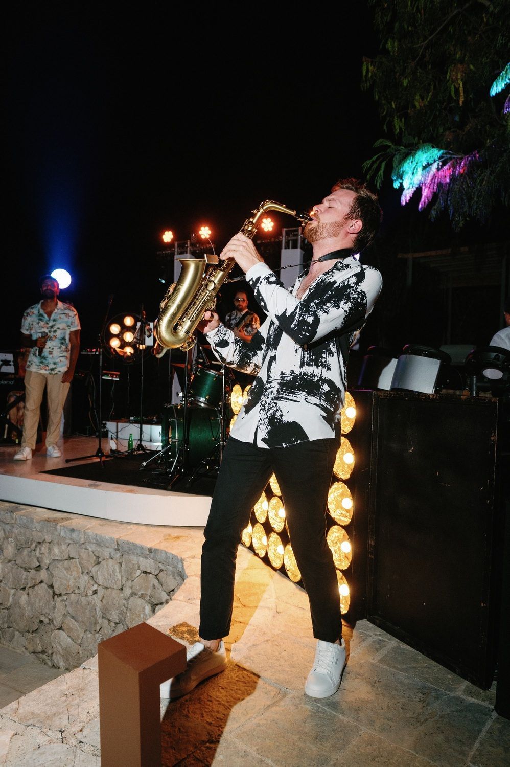 Saxophone player performs on an outdoor stone stage at night with warm lighting creating an atmospheric ambiance.