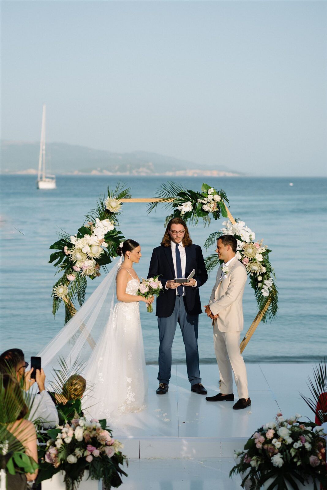 Beachfront wedding ceremony setup with white floral arch and ocean backdrop.