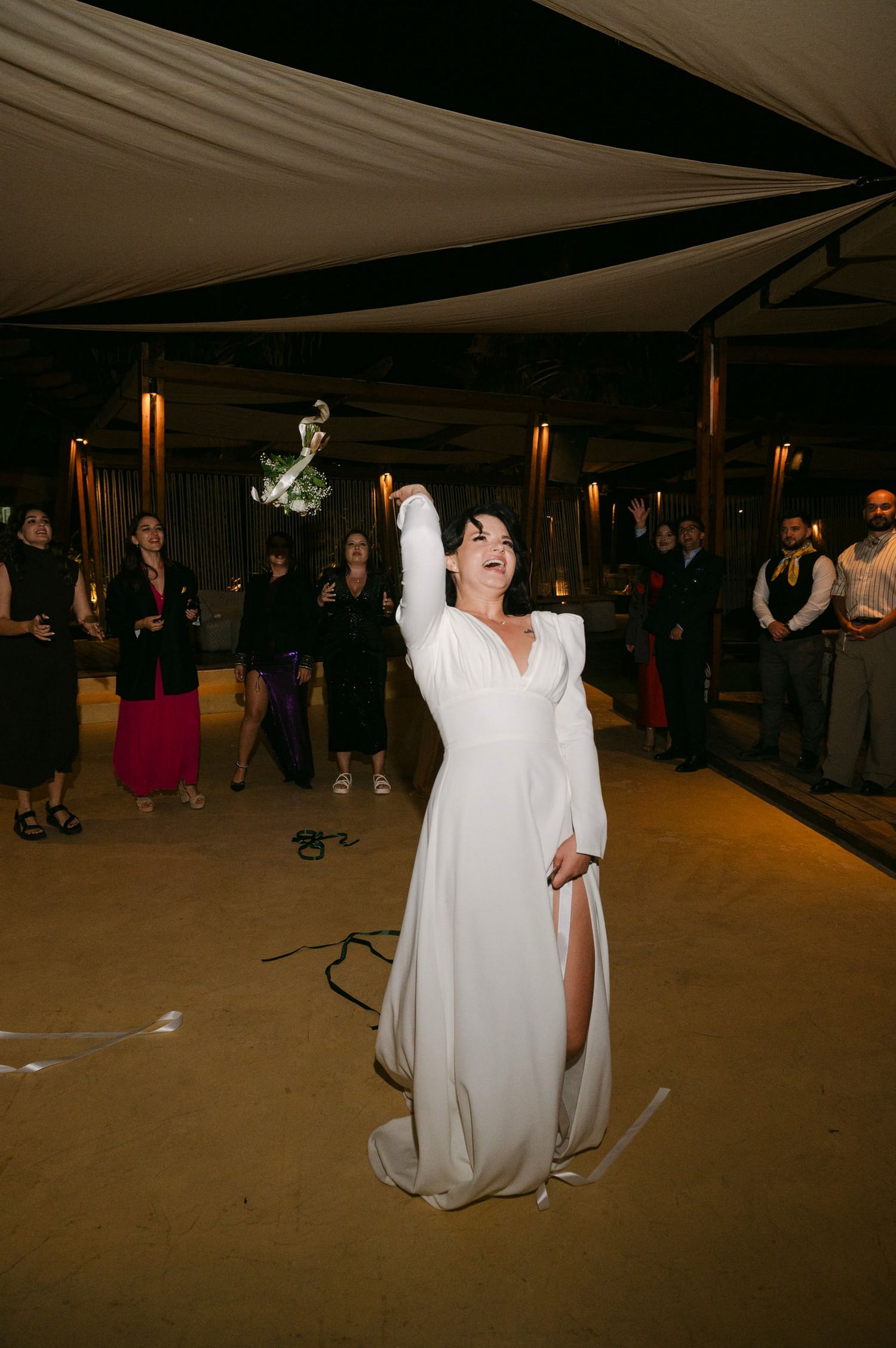 A group of people dance and celebrate at a dimly lit indoor event venue with white decor.