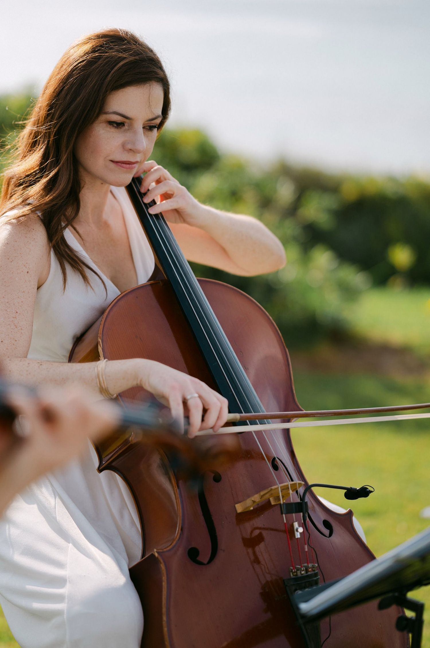 A cellist in a white dress plays their instrument outdoors in a sunny garden setting.