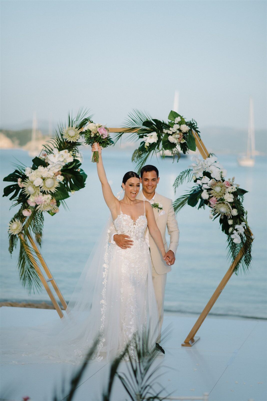 Beachside wedding ceremony setup with white floral arches and palm fronds against a serene ocean backdrop.