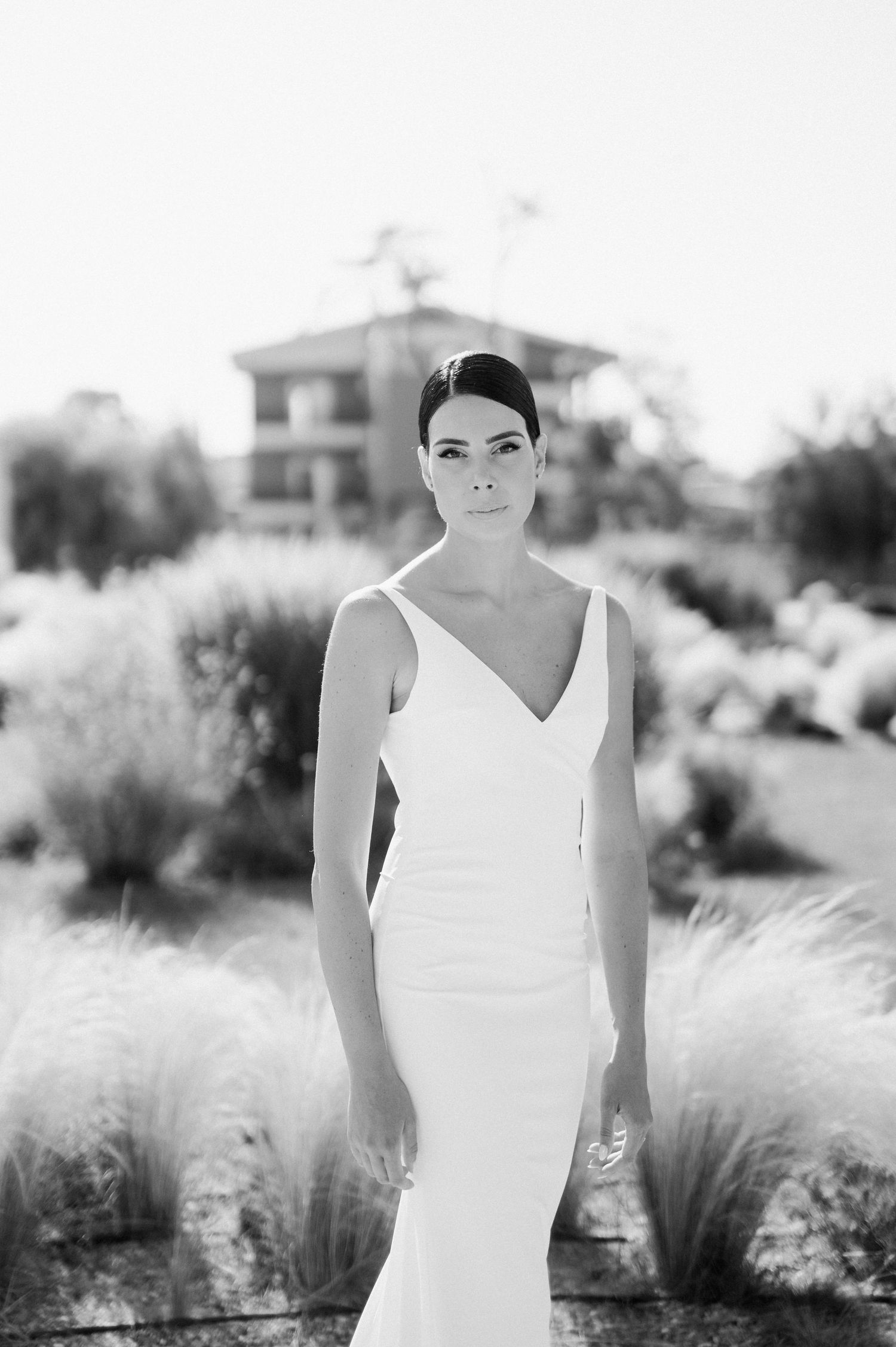 Black and white portrait of a bride in a fitted V-neck wedding dress standing in a garden with ornamental grasses.