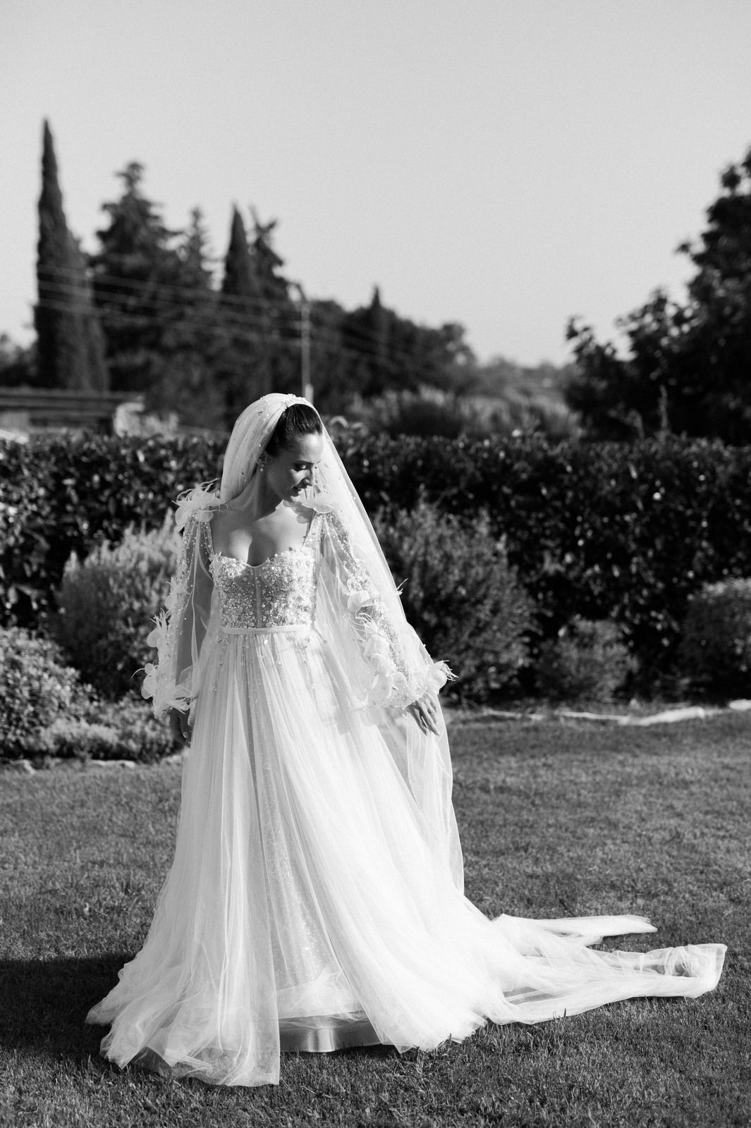 Elegant bride in flowing white lace gown and veil poses in garden setting.