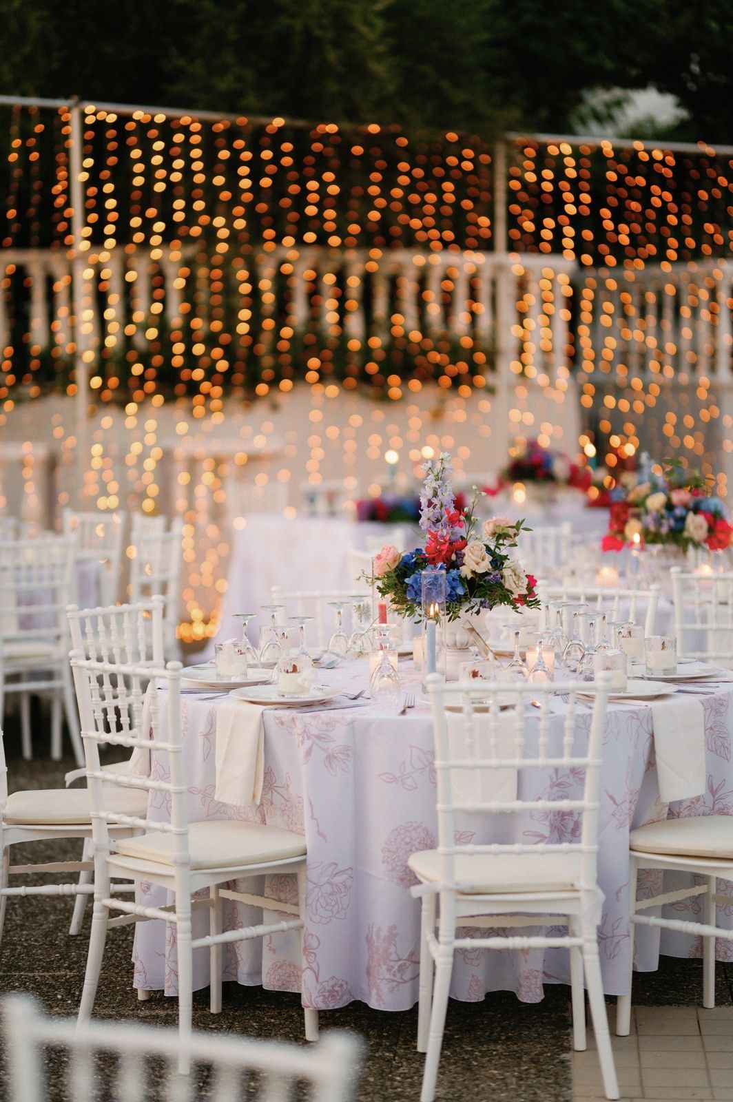 Elegant outdoor wedding reception setup with white chairs, fairy lights, and floral centerpieces on round tables.